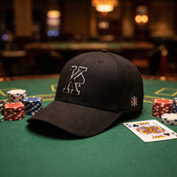Casquette baseball structurée noire brodée d'un grand K cerné de blanc avec un pique noir posée sur un tapis vert de table de poker à côté de jetons empilés et d'une carte de roi, lumière tamisée de casino - CHT'M Boutique