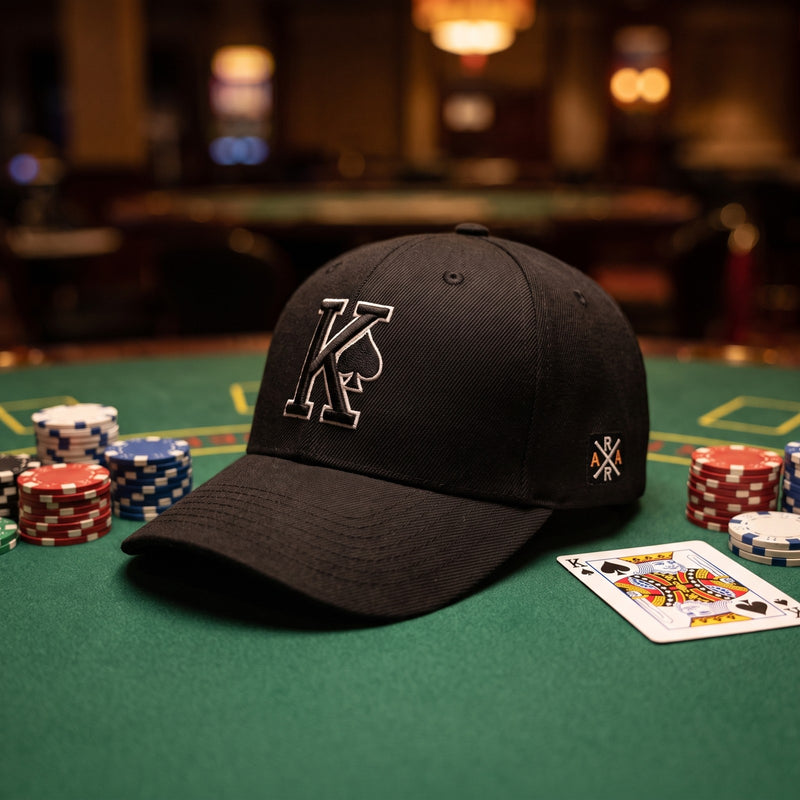 Casquette baseball structurée noire brodée d'un grand K cerné de blanc avec un pique noir posée sur un tapis vert de table de poker à côté de jetons empilés et d'une carte de roi, lumière tamisée de casino - CHT'M Boutique