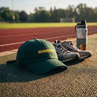 Casquette baseball vert brodée RUNGOOD en jaune posée sur une piste d'athlétisme à côté de chaussures de running et d'une gourde, lumière du petit matin - CHT'M Boutique