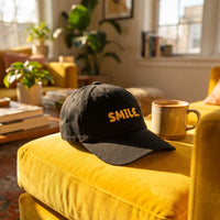 Casquette baseball noire brodée SMILE. en jaune posée sur un coussin jaune de canapé dans un salon ensoleillé à côté d'un mug, lumière joyeuse - CHT'M Boutique