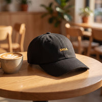 Casquette baseball noire brodée SMILE. en doré moutarde posée sur une table de café ensoleillée à côté d'un latte avec un coeur en mousse, lumière joyeuse du matin - CHT'M Boutique