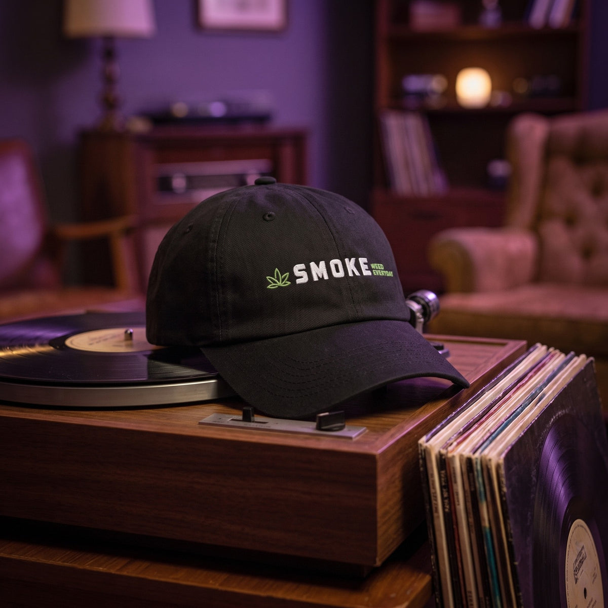 Casquette baseball noire brodée SMOKE en blanc avec une petite feuille verte et WEED EVERYDAY en vert posée sur une platine vinyle vintage à côté d'une pile de disques dans un salon chill tamisé avec une lumière violette - CHT'M Boutique
