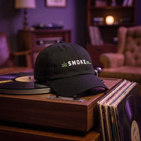 Casquette baseball noire brodée SMOKE en blanc avec une petite feuille verte et WEED EVERYDAY en vert posée sur une platine vinyle vintage à côté d'une pile de disques dans un salon chill tamisé avec une lumière violette - CHT'M Boutique