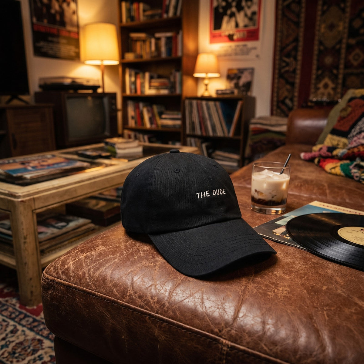 Casquette baseball noire brodée THE DUDE en blanc posée sur un canapé en cuir usé dans un salon des années 90 à côté d'un verre de white russian et d'un vinyle - CHT'M Boutique