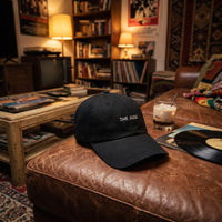 Casquette baseball noire brodée THE DUDE en blanc posée sur un canapé en cuir usé dans un salon des années 90 à côté d'un verre de white russian et d'un vinyle - CHT'M Boutique