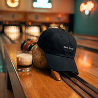 Casquette baseball noire brodée THE DUDE en blanc posée sur le retour de boules d'un bowling rétro à côté d'une boule et d'un verre de cocktail - CHT'M Boutique