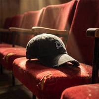 Casquette baseball noire effet usé brodée The End en écriture blanche, effet usé posée sur un siège de cinéma poussiéreux dans un théâtre abandonné au velours déchiré, lumière dorée poussiéreuse - CHT'M Boutique