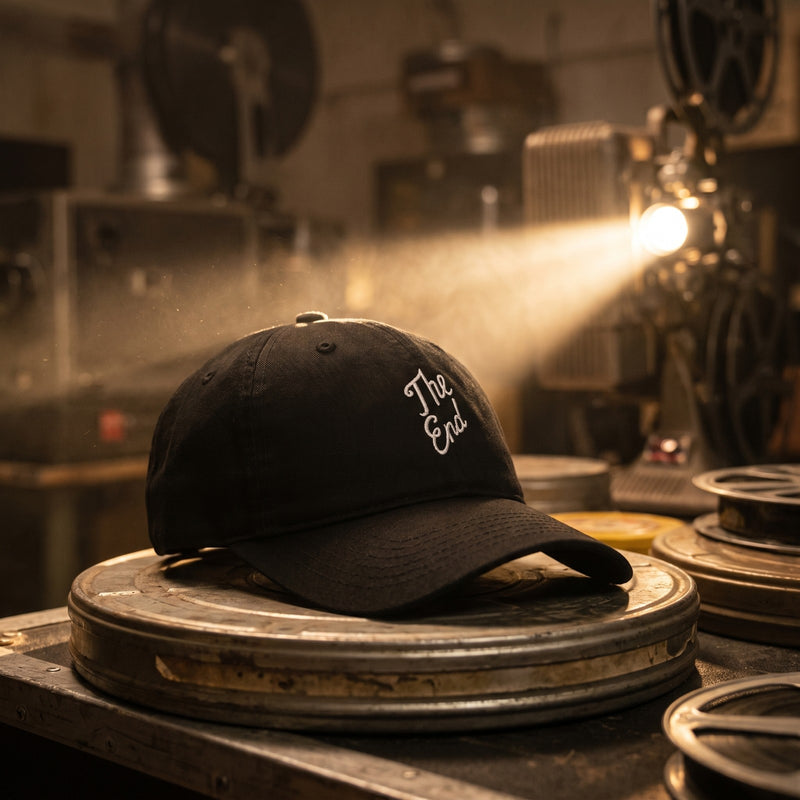Casquette baseball noire brodée The End en écriture blanche sur deux lignes posée sur une boîte de bobine de film dans une vieille cabine de projection avec un faisceau de lumière - CHT'M Boutique