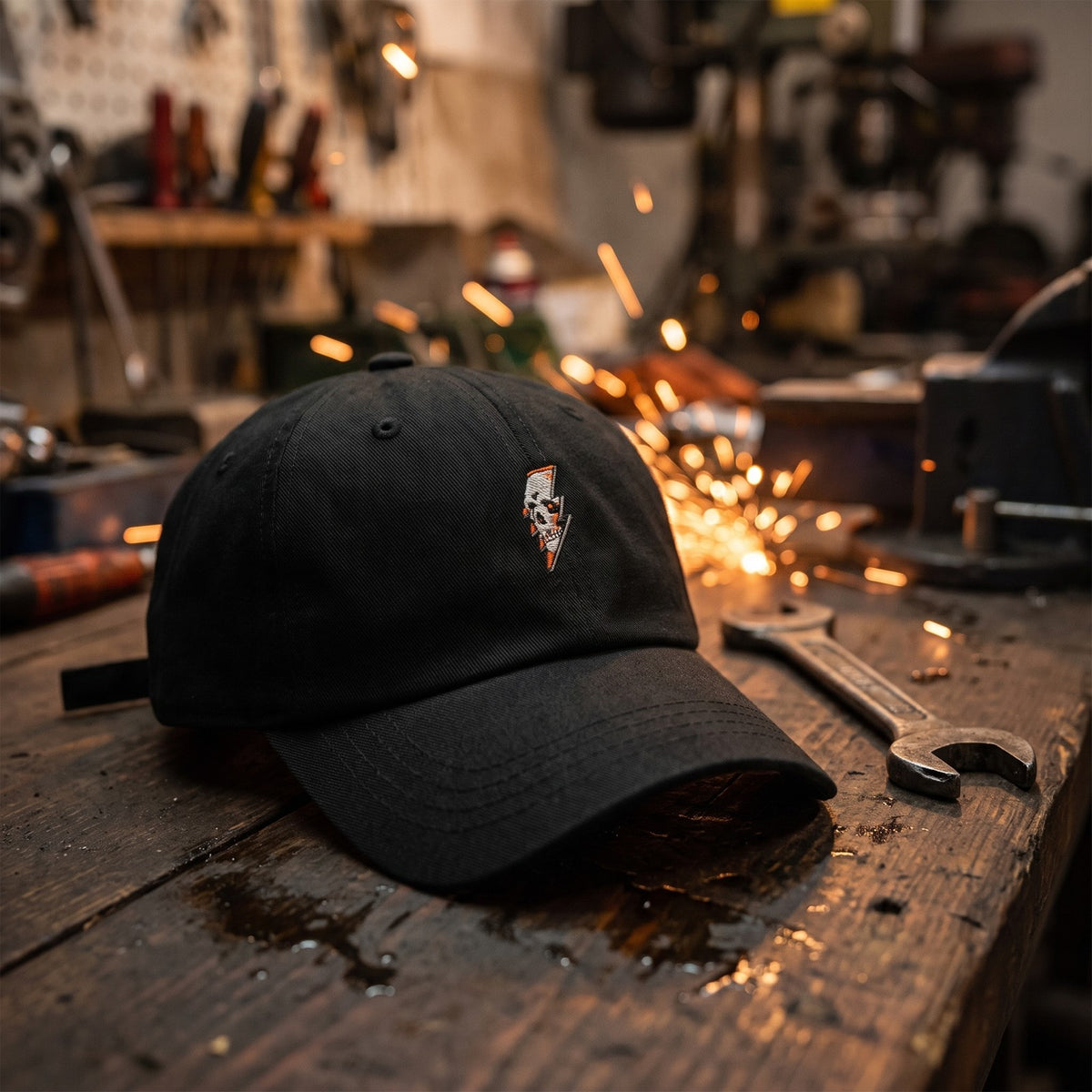 Casquette baseball noire brodée d'une petite tête de mort traversée par un éclair posée sur un établi d'atelier sombre à côté d'une clé à molette avec des étincelles orangées - CHT'M Boutique