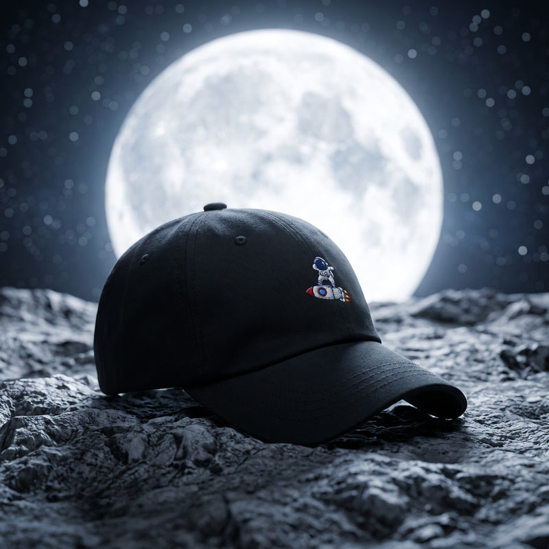 Casquette baseball noire brodée d'un petit astronaute avec le logo Cht'm posée sur une surface rocheuse lunaire devant une immense pleine lune et des étoiles, lumière nocturne cinématographique - CHT'M Boutique