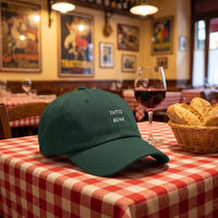 Casquette baseball vert épicéa brodée TUTTO BENE en blanc sur deux lignes posée sur une nappe à carreaux rouges dans une trattoria italienne à côté d'un verre de vin rouge et d'une corbeille de pain - CHT'M Boutique