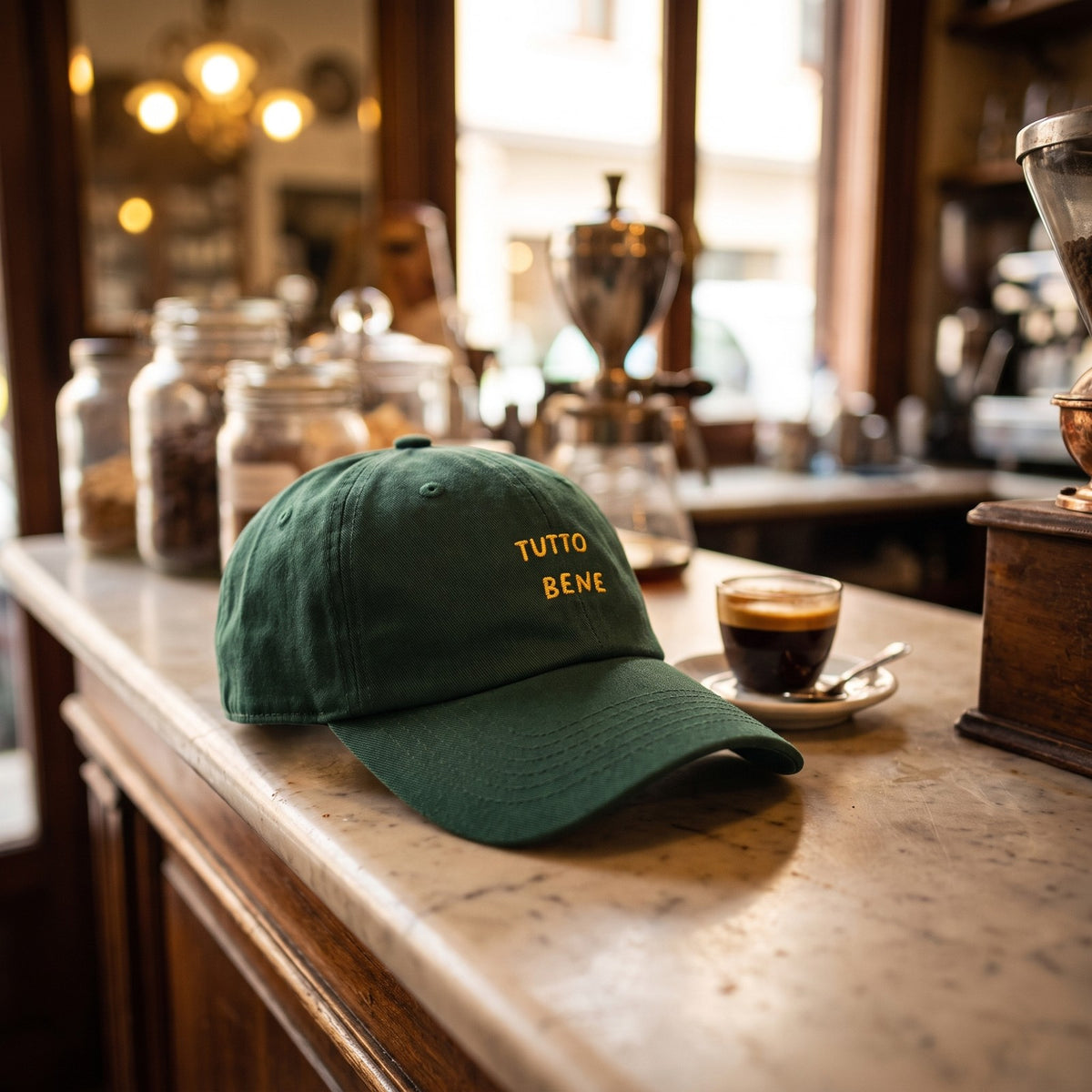 Casquette baseball vert épicéa brodée TUTTO BENE en jaune sur deux lignes posée sur le comptoir en marbre d'un bar à espresso italien à côté d'une tasse d'espresso - CHT'M Boutique