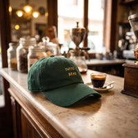 Casquette baseball vert épicéa brodée TUTTO BENE en jaune sur deux lignes posée sur le comptoir en marbre d'un bar à espresso italien à côté d'une tasse d'espresso - CHT'M Boutique