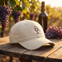 Casquette baseball oyster brodée d'un verre de vin bordeaux au trait posée sur une table rustique dans un vignoble avec du raisin et une bouteille sous le soleil chaud de fin de journée - CHT'M Boutique