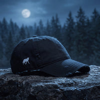 Casquette baseball noire effet usé brodée d'une petite silhouette de loup blanc sur le côté, effet usé posée sur une corniche rocheuse la nuit avec une lune pâle et une forêt de pins floue derrière, lumière bleue froide - CHT'M Boutique