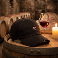 Casquette baseball noire brodée d'un petit verre de vin avec un coeur rouge posée sur un tonneau de vin dans une cave à côté d'un verre de vin rouge et d'une bougie - CHT'M Boutique