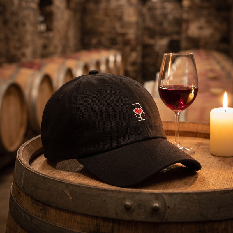Casquette baseball noire brodée d'un petit verre de vin avec un coeur rouge posée sur un tonneau de vin dans une cave à côté d'un verre de vin rouge et d'une bougie - CHT'M Boutique