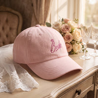 Casquette mariée rose pastel brodée Bride posée sur une coiffeuse de suite nuptiale avec un bouquet, un voile et des flûtes de champagne - CHT'M Boutique