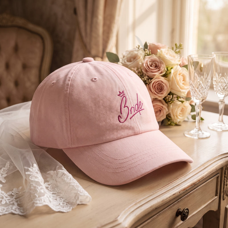 Casquette mariée rose pastel brodée Bride posée sur une coiffeuse de suite nuptiale avec un bouquet, un voile et des flûtes de champagne - CHT'M Boutique