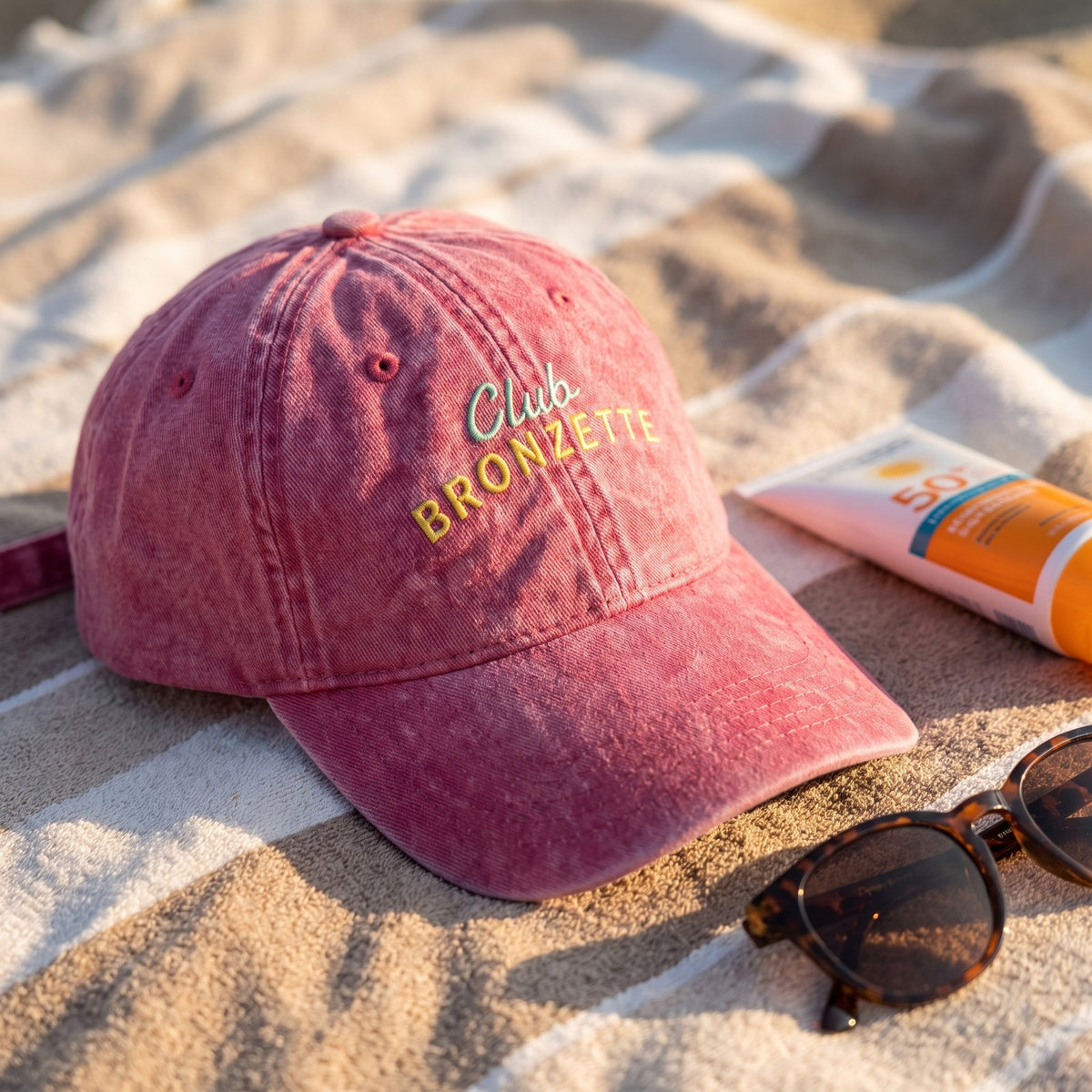 Casquette baseball rose Club Bronzette posée sur une serviette de plage à côté de crème solaire et de lunettes de soleil - CHT'M Boutique