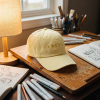 Casquette jaune pastel brodée Create posée sur la table à dessin d'un illustrateur avec des carnets et des feutres pastel - CHT'M Boutique