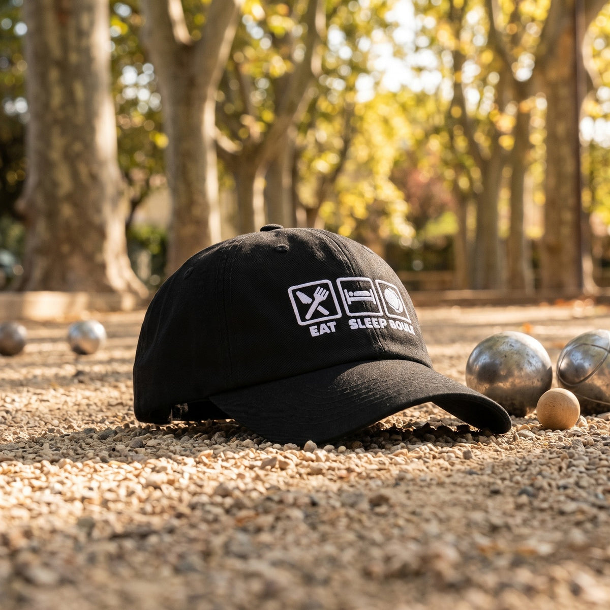 Casquette pétanque noire brodée EAT SLEEP BOULE posée sur le gravier d'un terrain de pétanque sous les platanes avec des boules - CHT'M Boutique