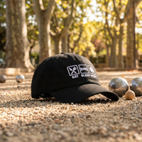 Casquette pétanque noire brodée EAT SLEEP BOULE posée sur le gravier d'un terrain de pétanque sous les platanes avec des boules - CHT'M Boutique