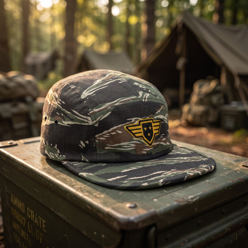 Casquette 5 panneaux camouflage tigre brodée d'un écusson étoile ailée jaune posée sur une caisse militaire olive patinée dans un camp en forêt à l'aube - CHT'M Boutique