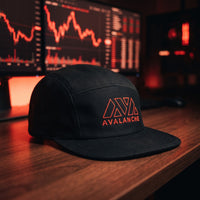 Casquette 5 panneaux noire brodée AVAX Avalanche en rouge posée sur un bureau de trader devant des écrans de graphiques rouges - CHT'M Boutique