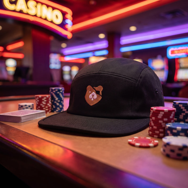 Casquette 5 panneaux noire brodée Bear Poker, ours marron avec cache-œil et pique posée sur un comptoir de casino avec des piles de jetons et un jeu de cartes - CHT'M Boutique