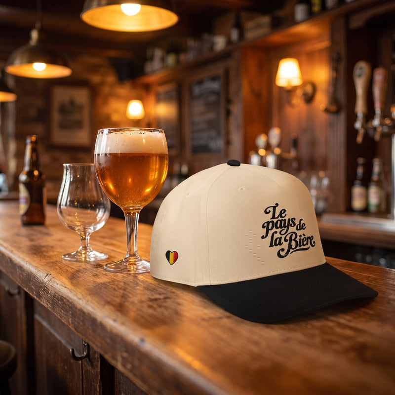 Casquette 5 panneaux crème et noire brodée « Le pays de la Bière » posée sur le comptoir en bois d'un bar à bière belge à côté de verres de bière - CHT'M Boutique