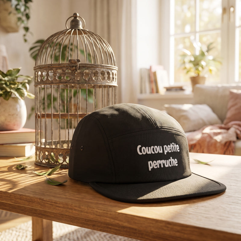 Casquette 5 panneaux noire brodée Coucou petite perruche posée près d'une cage à oiseaux ouvragée dans un salon lumineux avec des plumes vertes - CHT'M Boutique