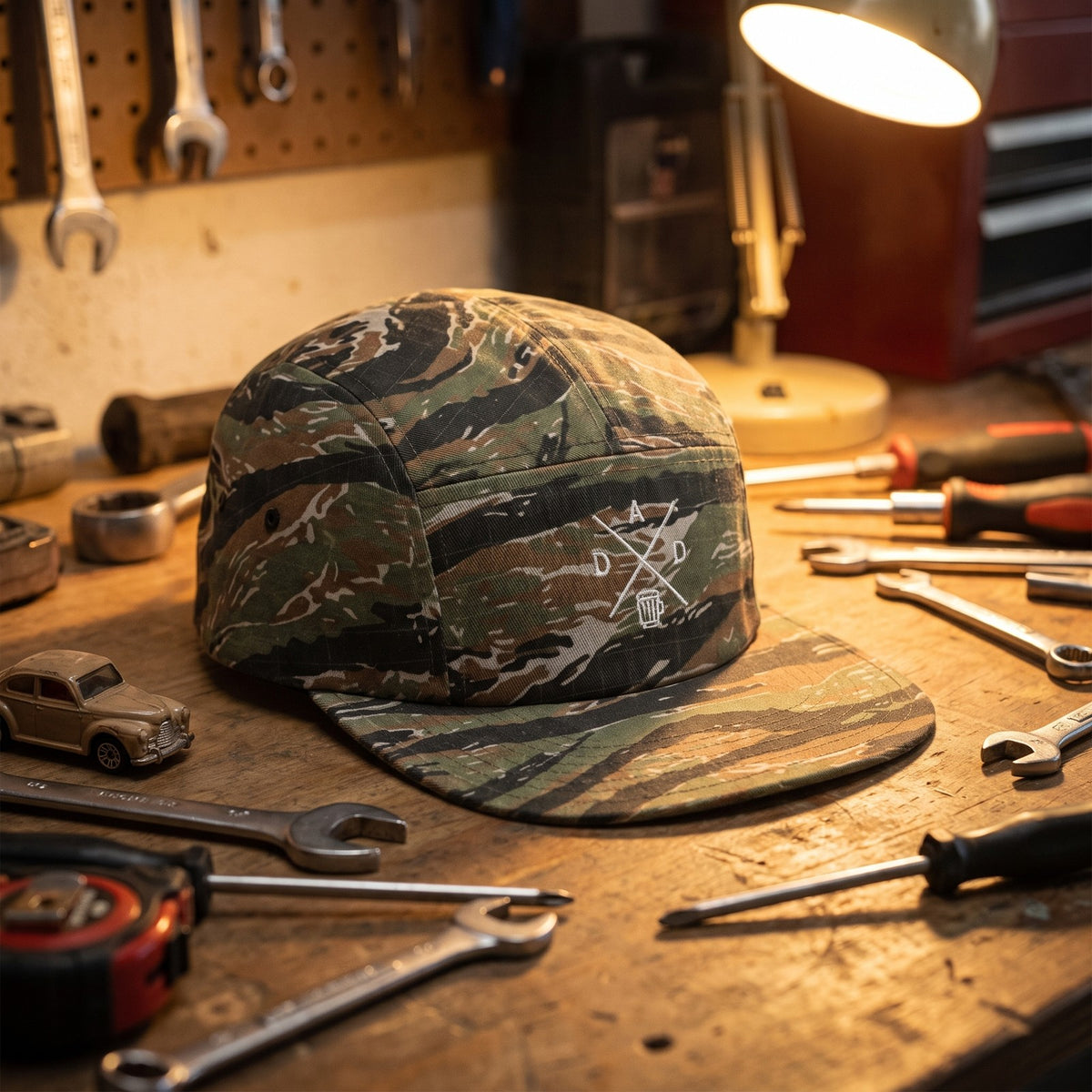 Casquette 5 panneaux camouflage tigre brodée du monogramme DAD blanc posée sur un établi de garage avec des outils et une petite voiture - CHT'M Boutique