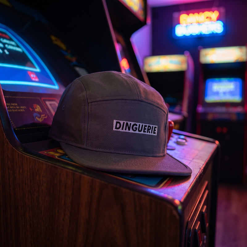 Casquette 5 panneaux gris anthracite brodée DINGUERIE posée sur une borne d'arcade rétro dans une salle de jeux éclairée au néon - CHT'M Boutique
