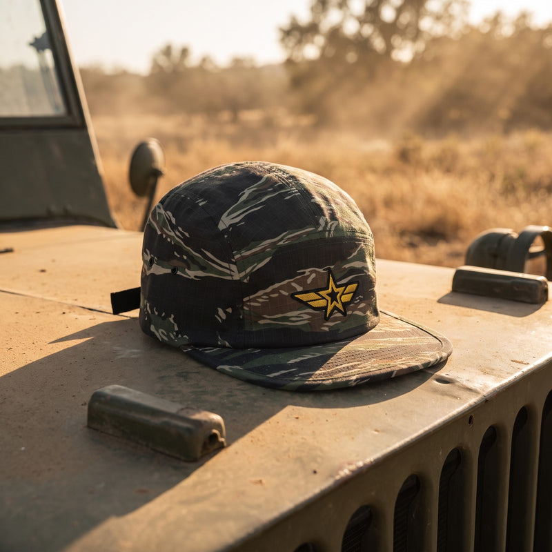 Casquette militaire 5 panneaux camouflage tigre brodée d'une étoile ailée jaune posée sur le capot d'une jeep militaire vintage dans un champ poussiéreux - CHT'M Boutique