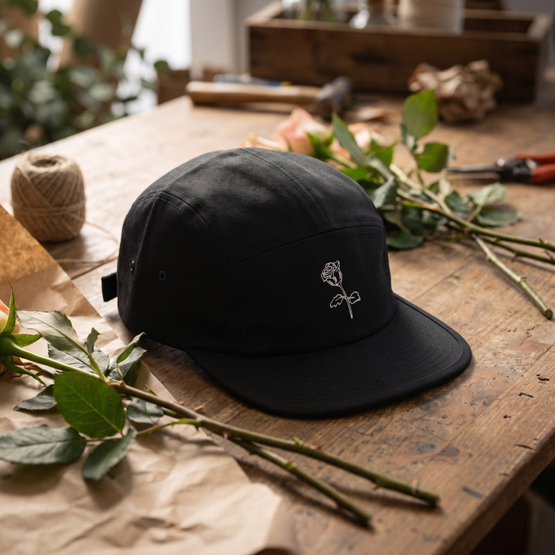Casquette 5 panneaux noire brodée d'une rose blanche au trait posée sur l'établi d'un fleuriste avec des tiges de roses et du papier kraft - CHT'M Boutique