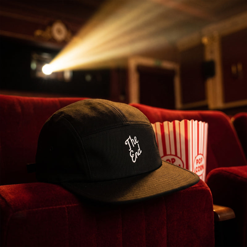 Casquette 5 panneaux noire brodée The End en écriture manuscrite blanche posée sur le fauteuil en velours d'un cinéma vintage vide avec un pot de pop-corn - CHT'M Boutique