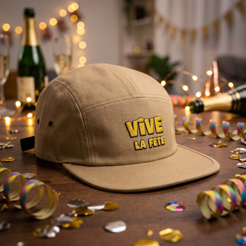 Casquette 5 panneaux kaki brodée Vive la fête en jaune posée sur une table couverte de confettis avec des serpentins et une bouteille de champagne - CHT'M Boutique