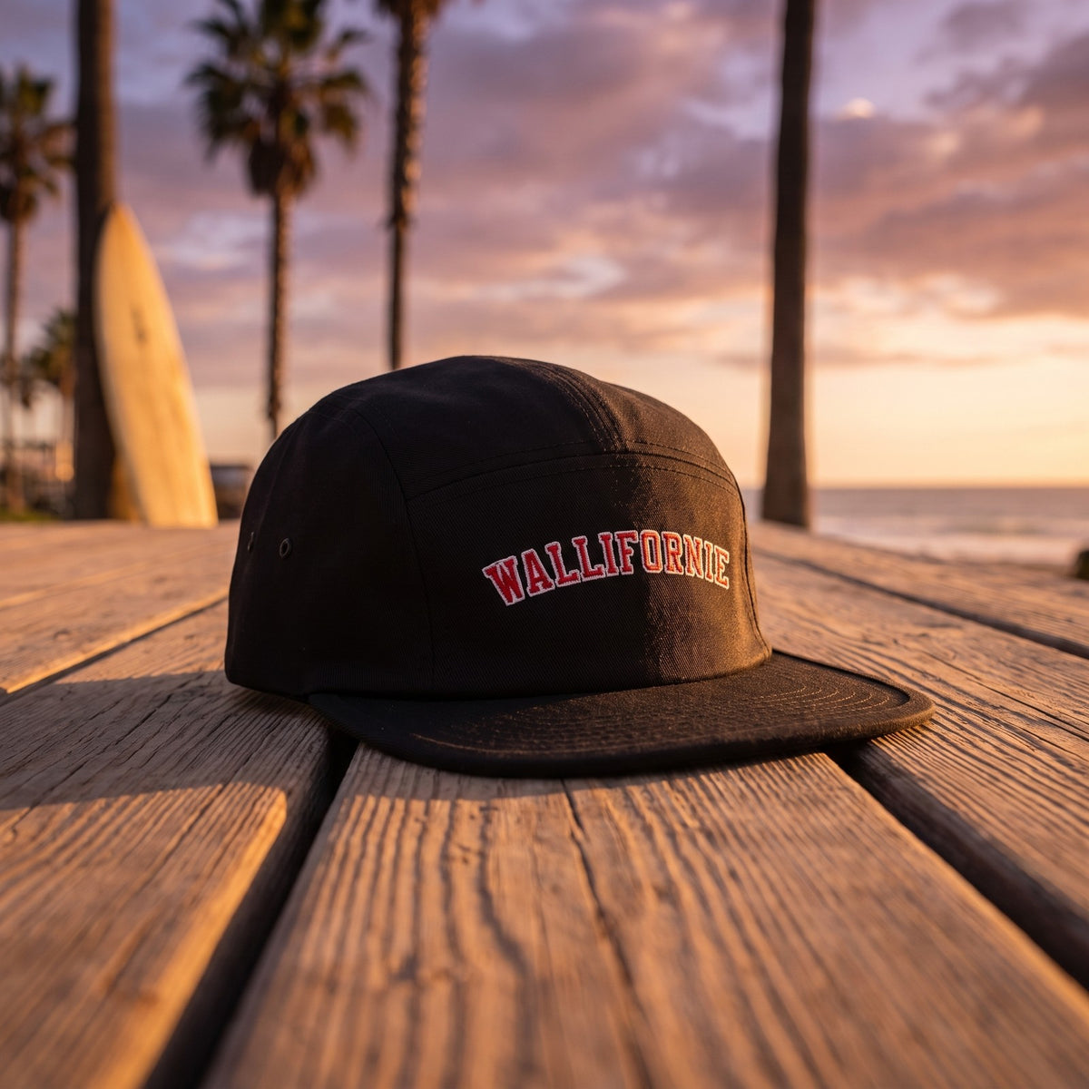 Casquette 5 panneaux noire brodée WALLIFORNIE en lettres universitaires rouges et blanches posée sur une promenade en bois avec des palmiers et une planche de surf au coucher du soleil - CHT'M Boutique