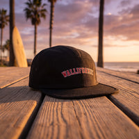 Casquette 5 panneaux noire brodée WALLIFORNIE en lettres universitaires rouges et blanches posée sur une promenade en bois avec des palmiers et une planche de surf au coucher du soleil - CHT'M Boutique