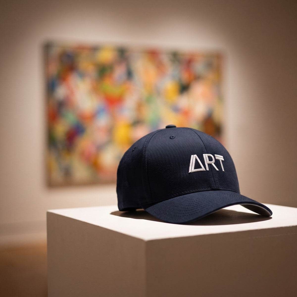 Casquette Flexfit fermée à l'arrière bleu marine brodée ART en blanc posée sur un socle blanc dans une galerie d'art moderne devant un tableau abstrait flou - CHT'M Boutique