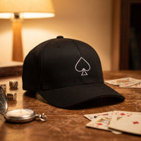 Casquette Flexfit fermée à l'arrière noire brodée d'un as de pique blanc au trait, Play Time posée sur une table de jeu vintage à côté de dés, d'une montre à gousset et de cartes éparpillées - CHT'M Boutique