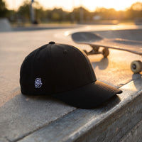Casquette Flexfit fermée à l'arrière noire avec petit logo CHT'M brodé en blanc sur le côté posée sur un muret en béton d'un skatepark à côté d'un skateboard au coucher du soleil - CHT'M Boutique
