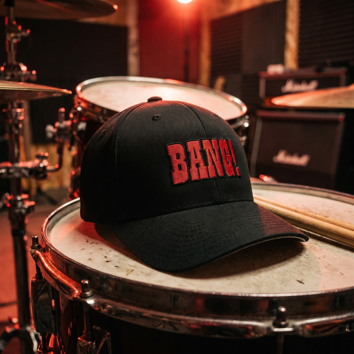 Casquette Flexfit fermée à l'arrière noire brodée BANG! en rouge posée sur une caisse claire dans une salle de répétition avec des baguettes à côté - CHT'M Boutique