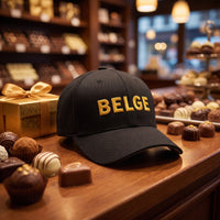 Casquette Flexfit noire brodée BELGE dorée posée sur le comptoir d'un chocolatier belge à côté de pralines et d'une boîte dorée - CHT'M Boutique
