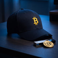 Casquette Flexfit fermée à l'arrière bleu marine brodée du symbole Bitcoin doré en 3D puff posée sur un bureau minimaliste à côté d'un portefeuille crypto et d'une pièce dorée - CHT'M Boutique