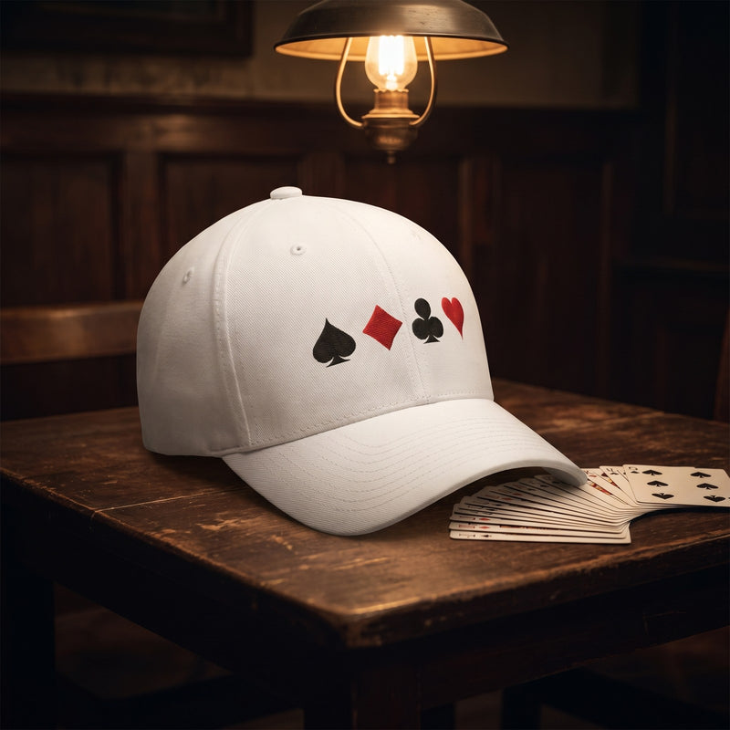 Casquette Flexfit fermée à l'arrière blanche brodée des quatre couleurs d'un jeu de cartes posée sur une table de cartes en bois dans une arrière-salle à côté d'un éventail de cartes sous une lampe suspendue - CHT'M Boutique