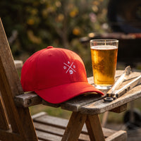 Casquette Flexfit fermée à l'arrière rouge brodée du monogramme DAD blanc avec une chope de bière posée sur l'accoudoir en bois d'un transat de jardin à côté d'une bière fraîche et de pinces de barbecue - CHT'M Boutique