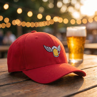 Casquette Flexfit fermée à l'arrière rouge brodée d'une chope de bière dorée avec des ailes posée sur une table en bois d'un jardin à bière d'été avec des guirlandes lumineuses et une bière mousseuse - CHT'M Boutique