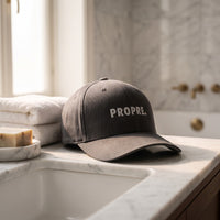 Casquette Flexfit fermée à l'arrière gris anthracite brodée PROPRE. en gris clair posée sur le rebord d'un lavabo en marbre blanc impeccable à côté de serviettes blanches pliées et d'un savon - CHT'M Boutique